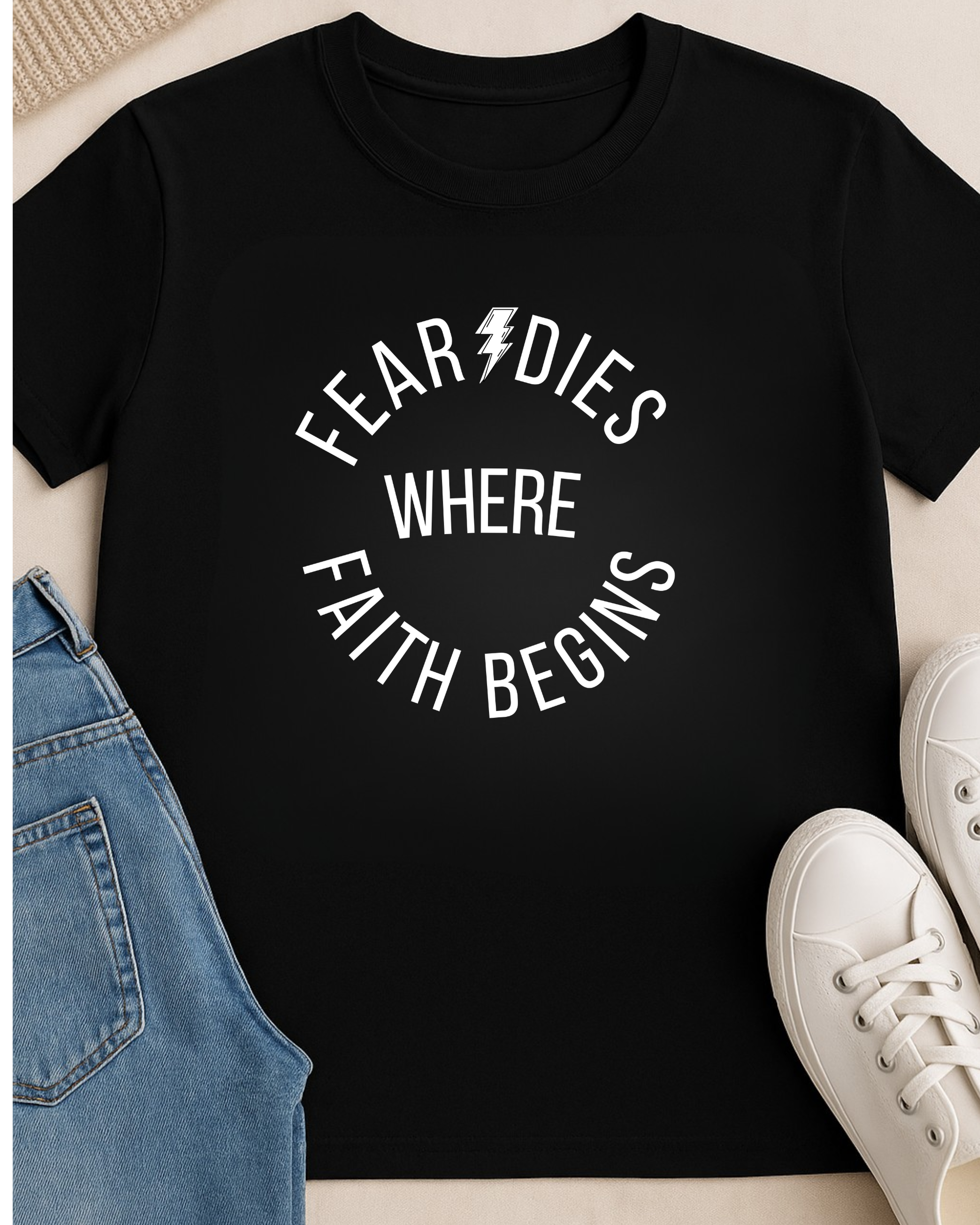 Flat lay of “Fear Dies Where Faith Begins” black t-shirt styled with jeans and white sneakers
