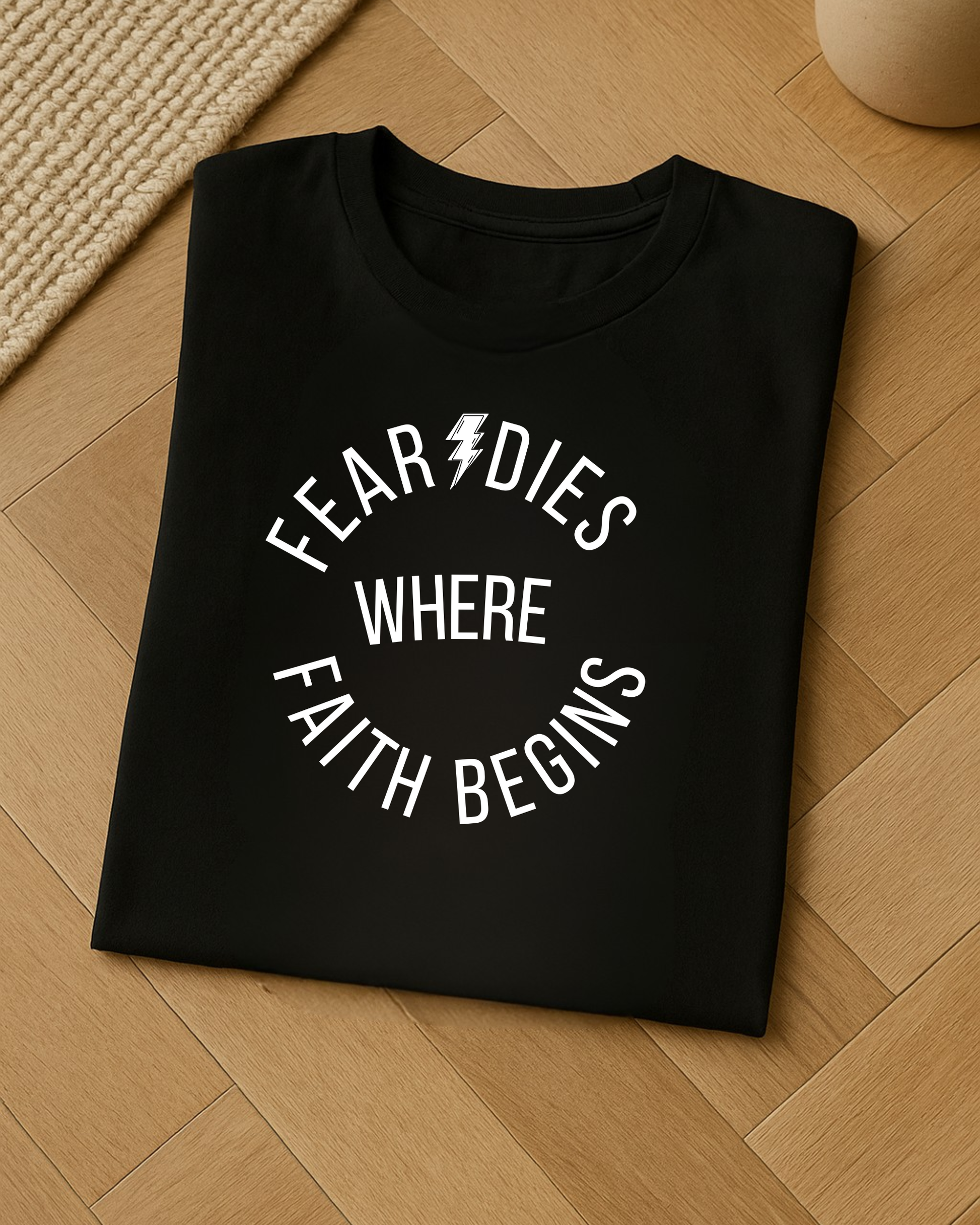Folded black t-shirt with “Fear Dies Where Faith Begins” design on wooden floor