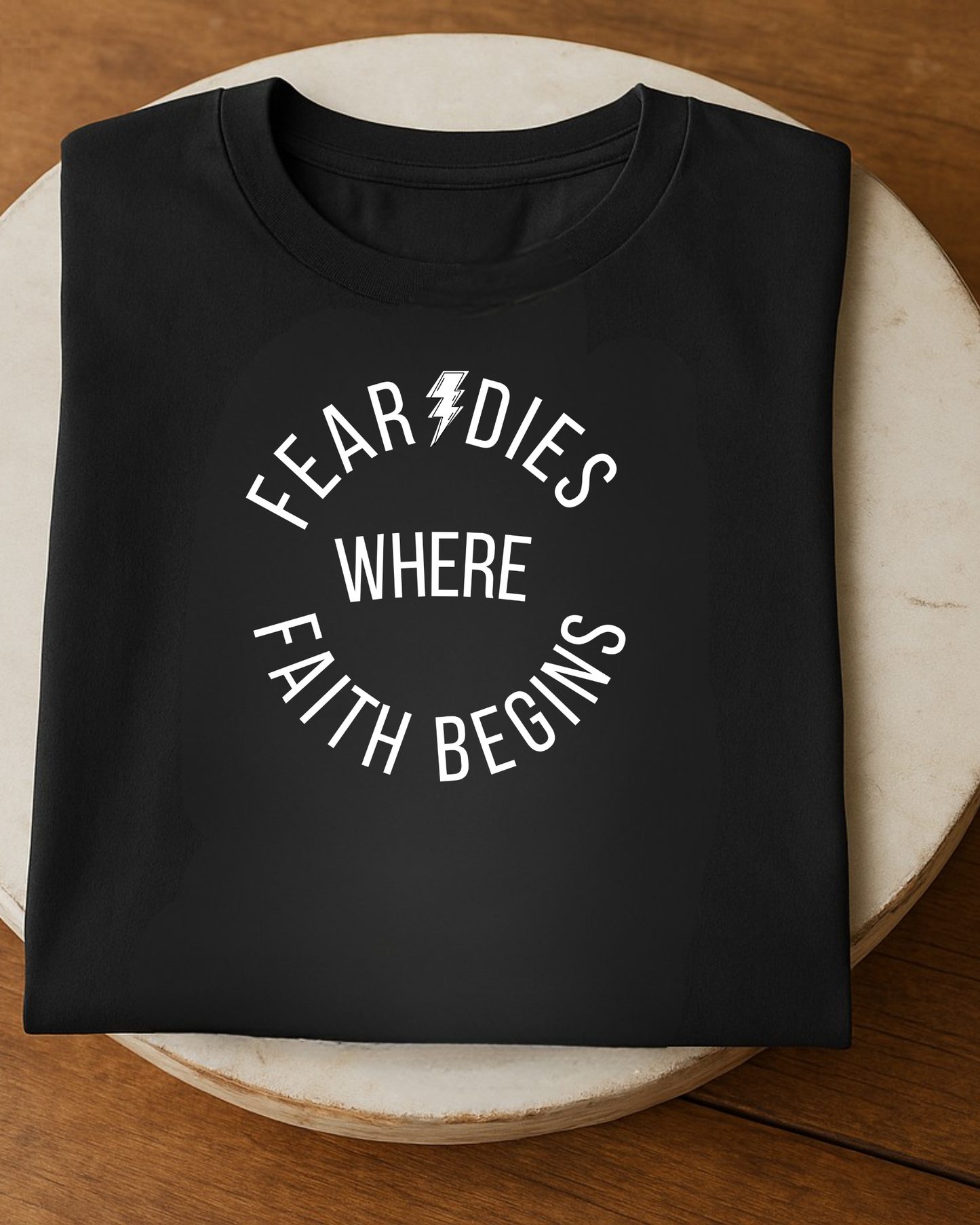 Folded black t-shirt with “Fear Dies Where Faith Begins” design on round wooden table