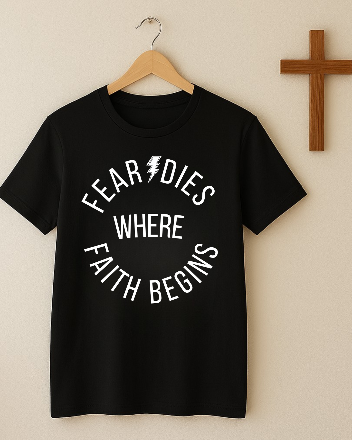 Hanging black Christian t-shirt with “Fear Dies Where Faith Begins” beside wooden cross