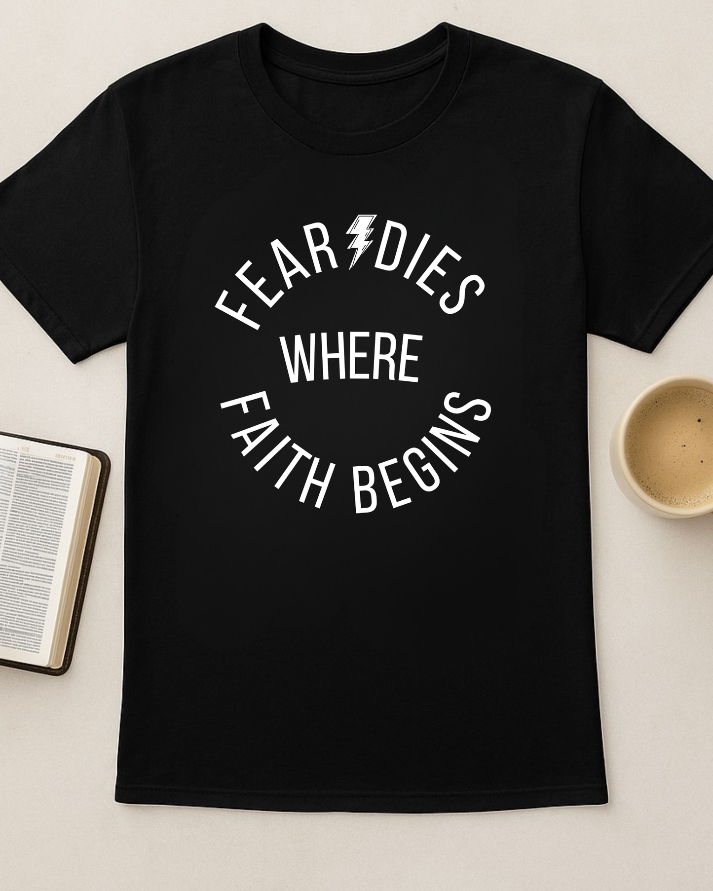 Black “Fear Dies Where Faith Begins” t-shirt laid out with open Bible and coffee cup