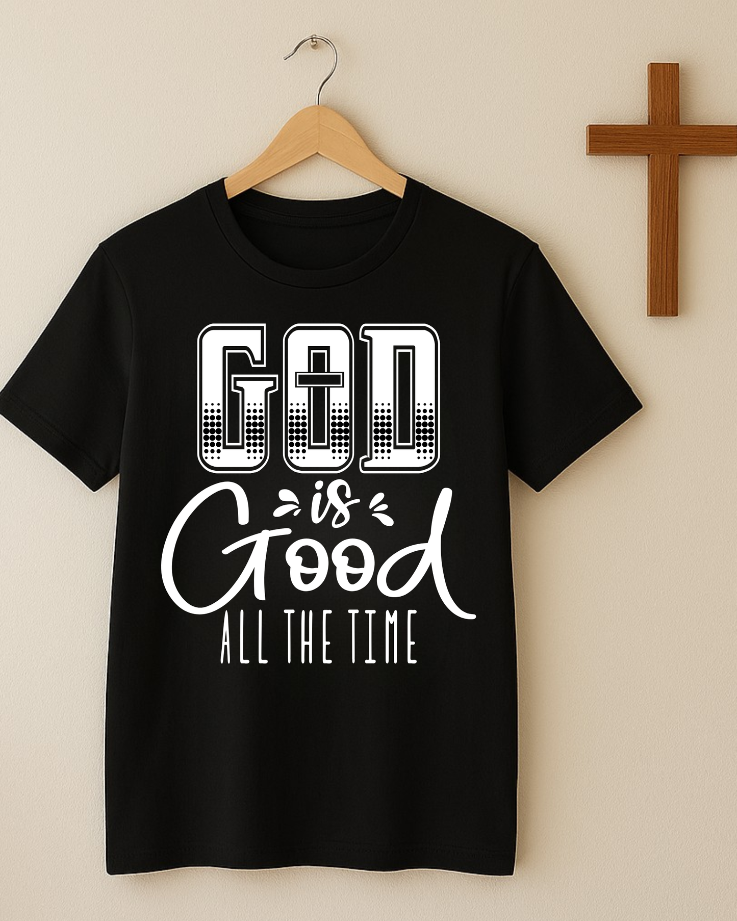 Black Christian God Is Good All The Time t-shirt hanging on a wooden hanger next to a cross on a beige wall.