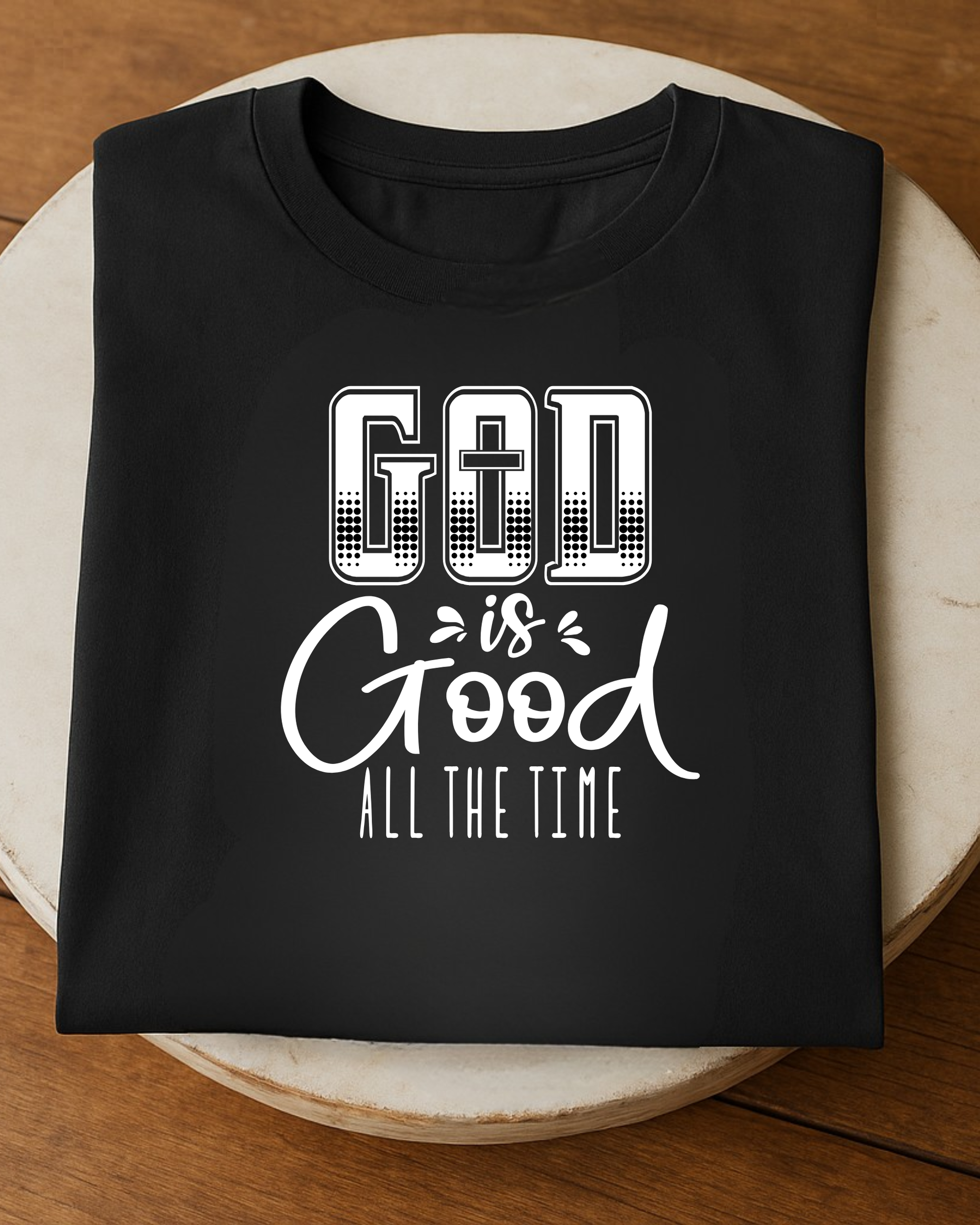 Folded black Christian God Is Good All The Time t-shirt displayed on a round wooden table.