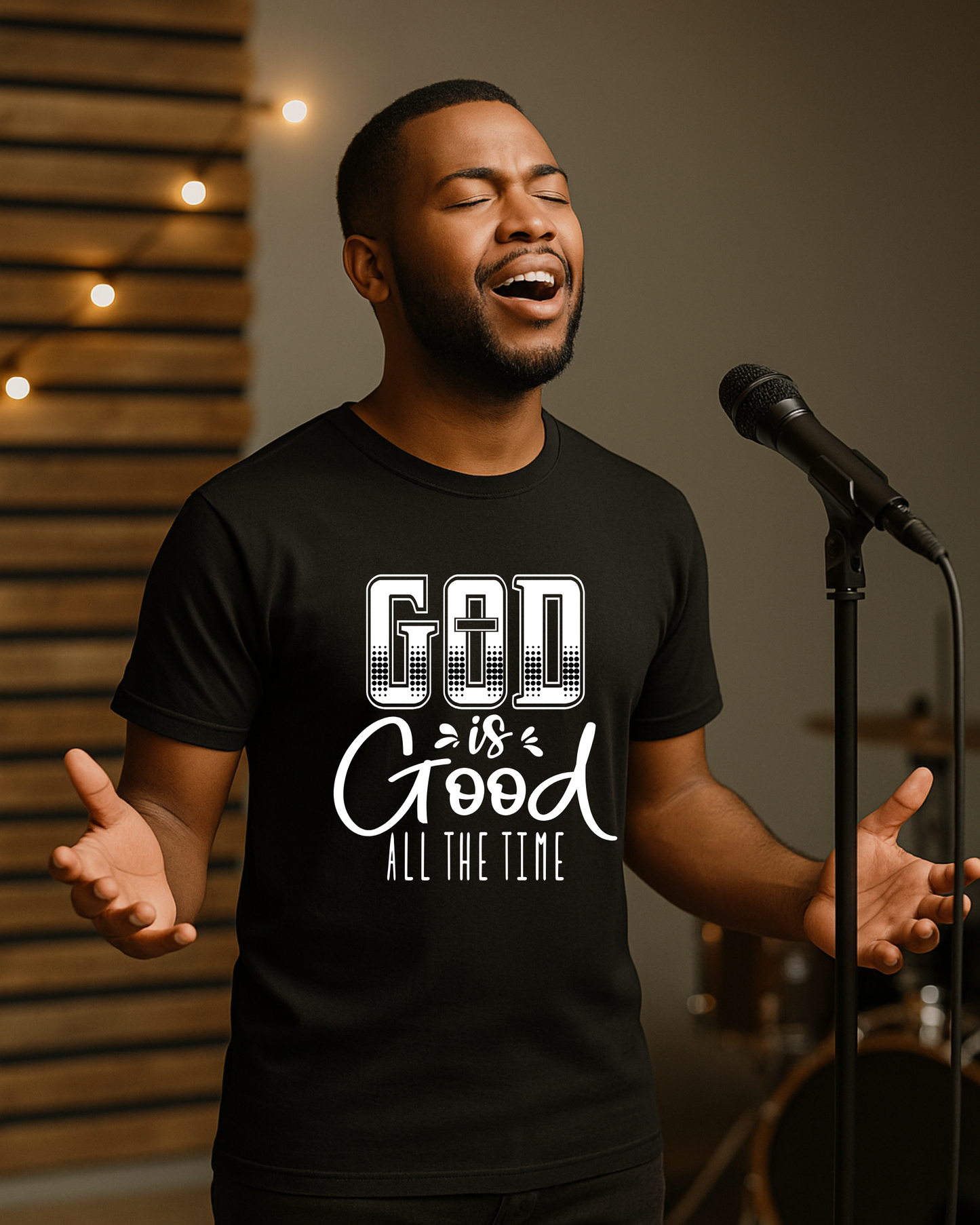 Christian man singing in worship while wearing a black God Is Good All The Time t-shirt on stage with a microphone.