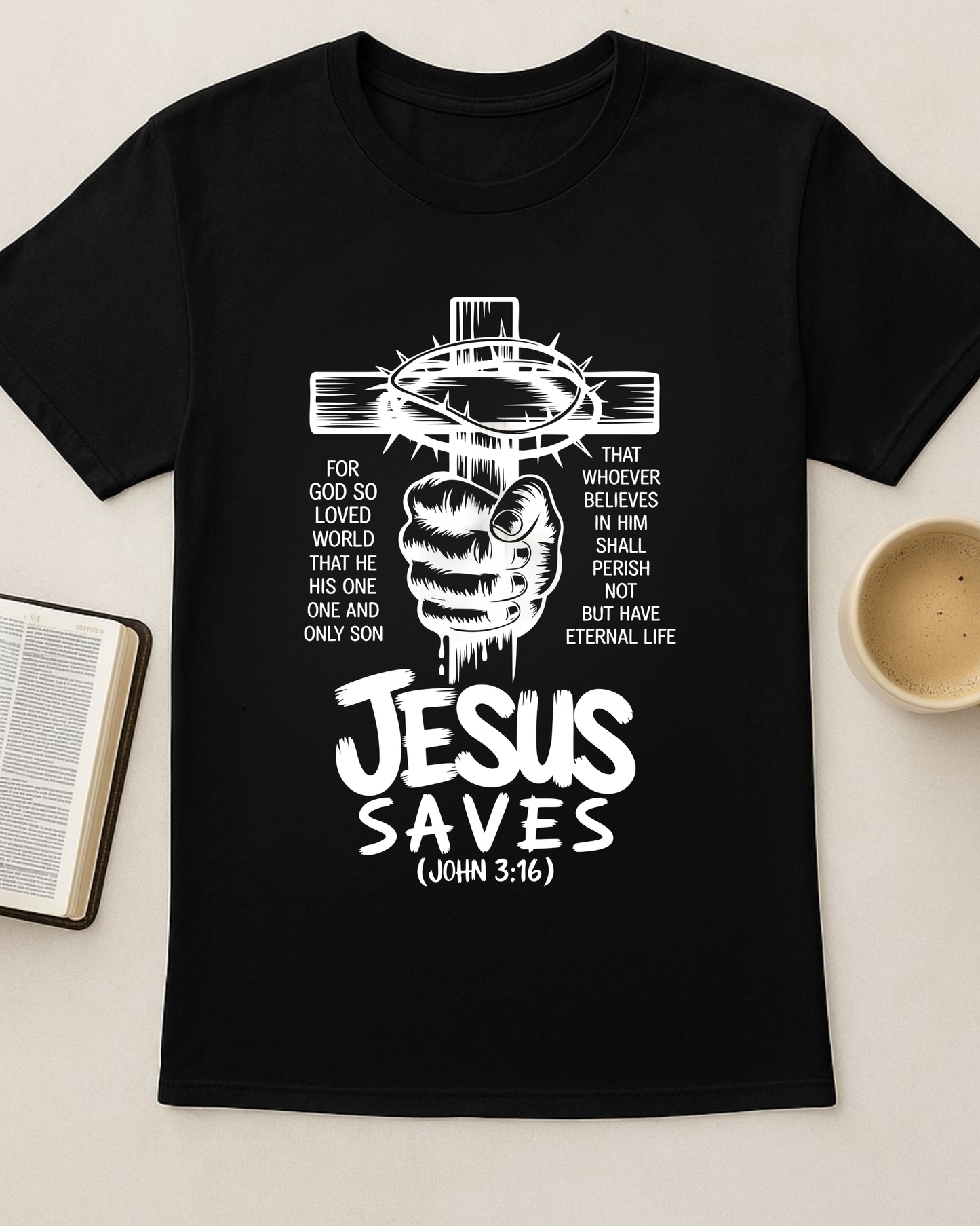 Black Christian T-shirt with “Jesus Saves John 3:16” cross design displayed flat beside an open Bible and a cup of coffee.