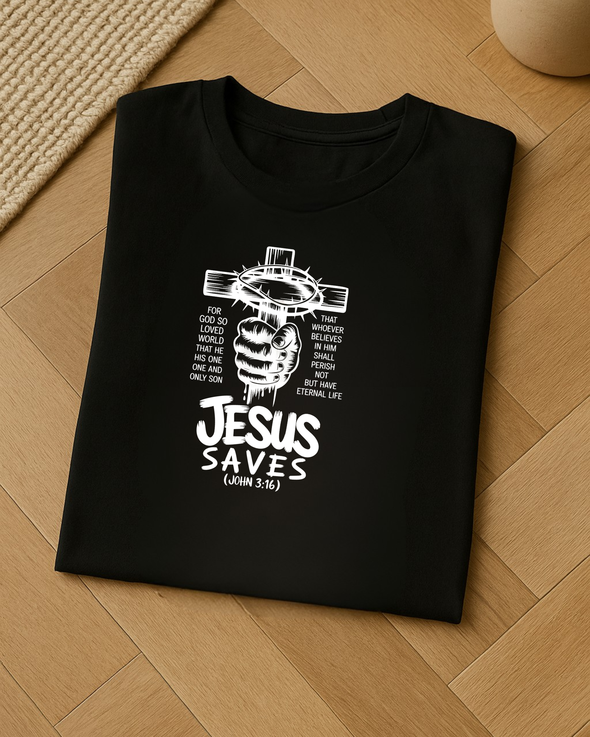 Folded black Christian T-shirt featuring a “Jesus Saves John 3:16” cross design on a wooden floor background.