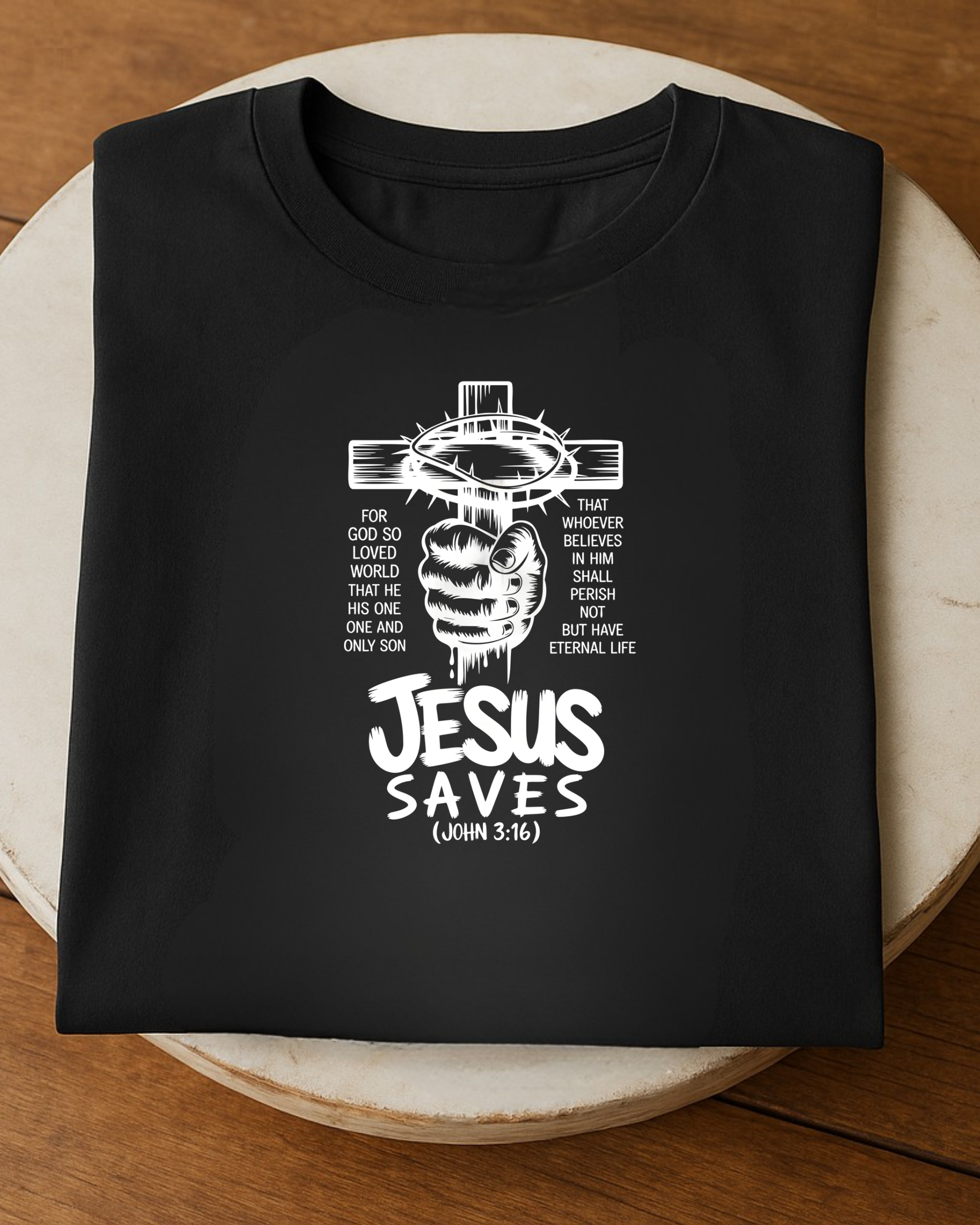 Folded black Christian T-shirt with “Jesus Saves John 3:16” cross design displayed on a round wooden table.