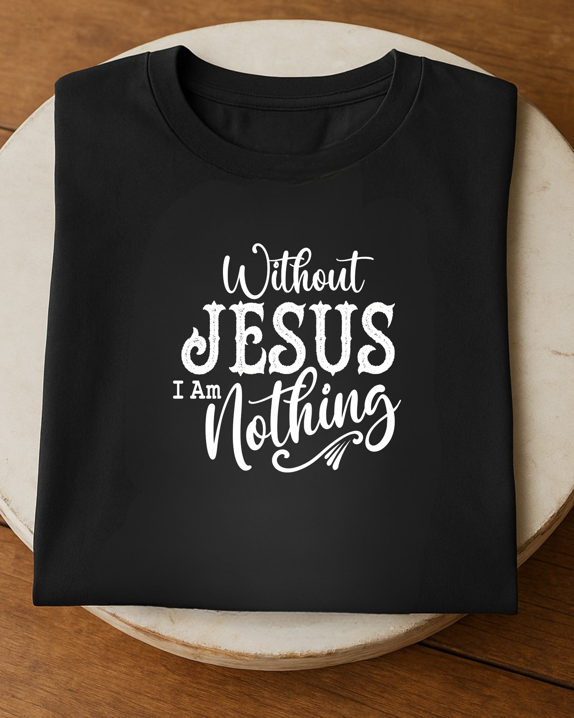 Black Christian T-shirt folded on a round wooden table featuring the quote “Without Jesus I Am Nothing” in white decorative lettering.