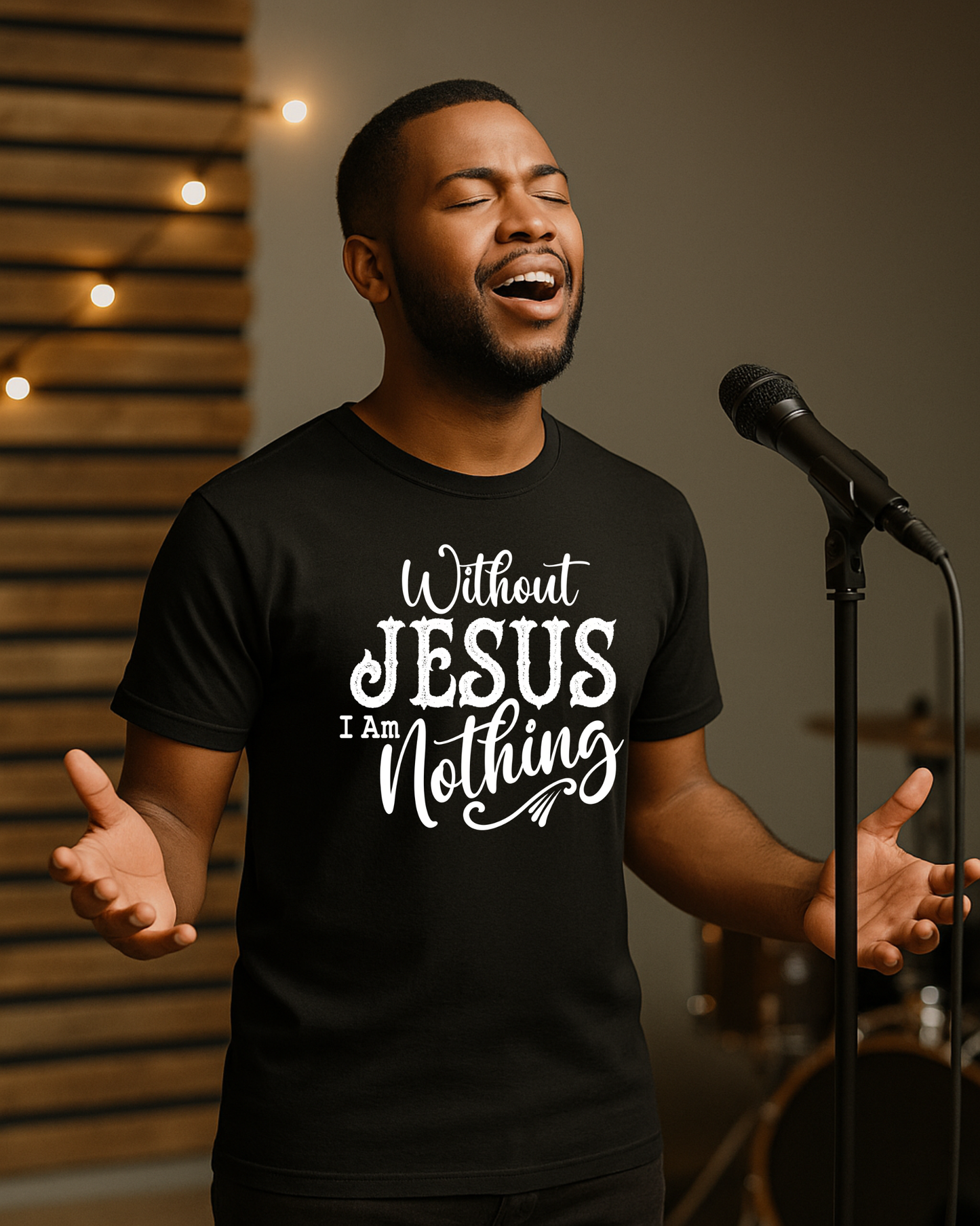 Christian worship leader singing on stage wearing a black T-shirt that reads “Without Jesus I Am Nothing” with microphone and warm studio lighting in the background.